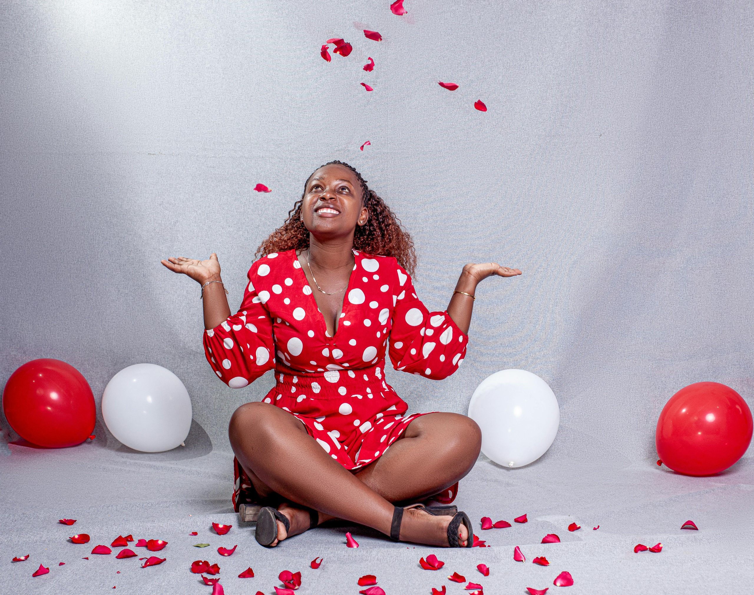 Smiling woman in red polka dot dress surrounded by red and white balloons indoors.