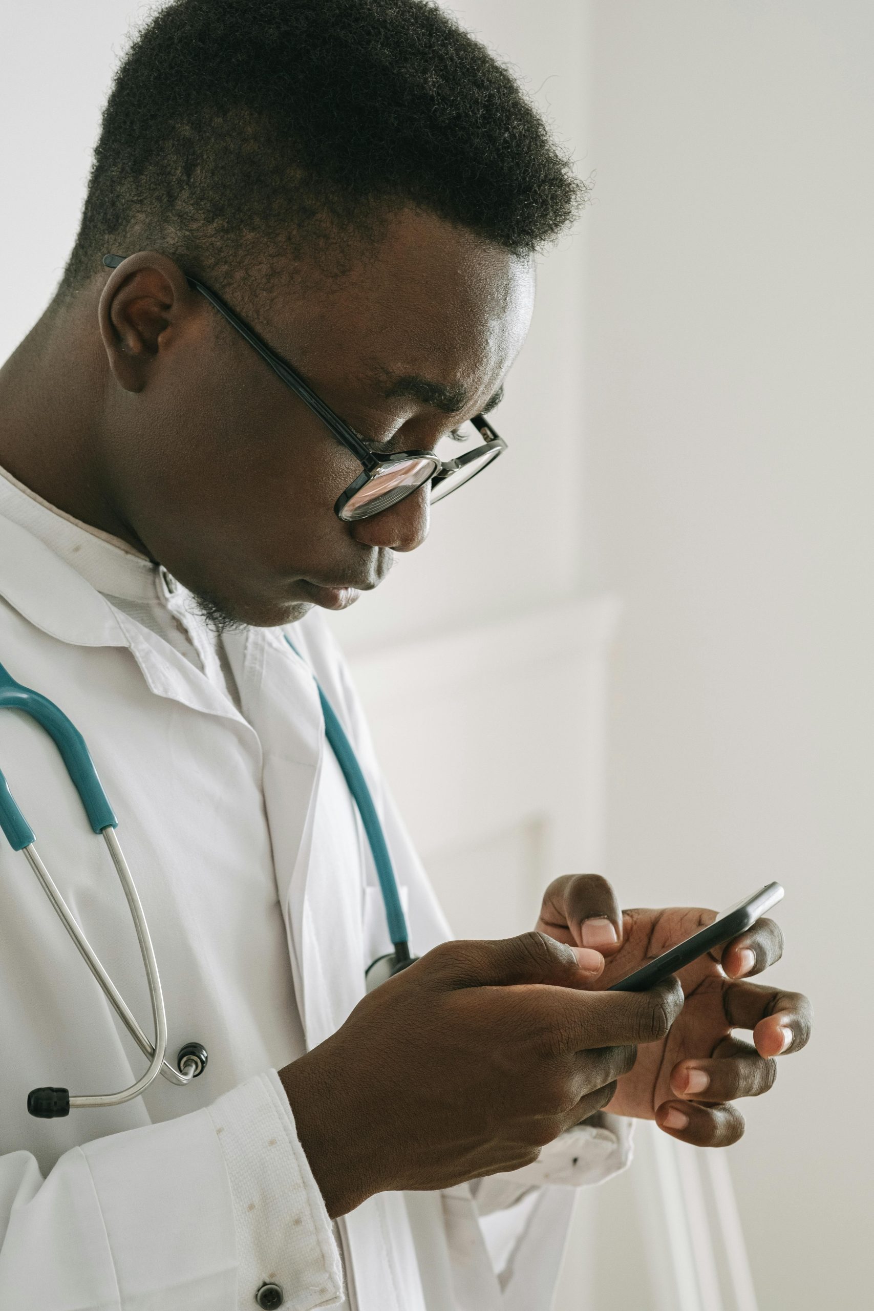 African American doctor in a lab coat checks his smartphone while standing indoors.
